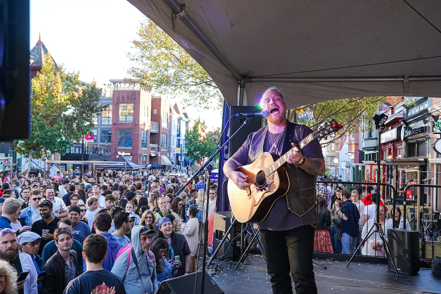 A musician passionately performs on stage during Adams Morgan PorchFest in Washington, DC, with a vibrant crowd behind him.