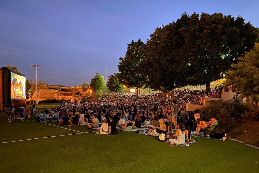A large crowd watches an outdoor movie screening at night, with trees and city lights in the background.